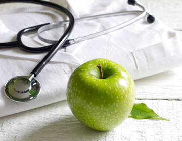 stetoskop lekarski na białym fartuchu lekarskim oraz zielone jabłko symbolizujące zdrowie